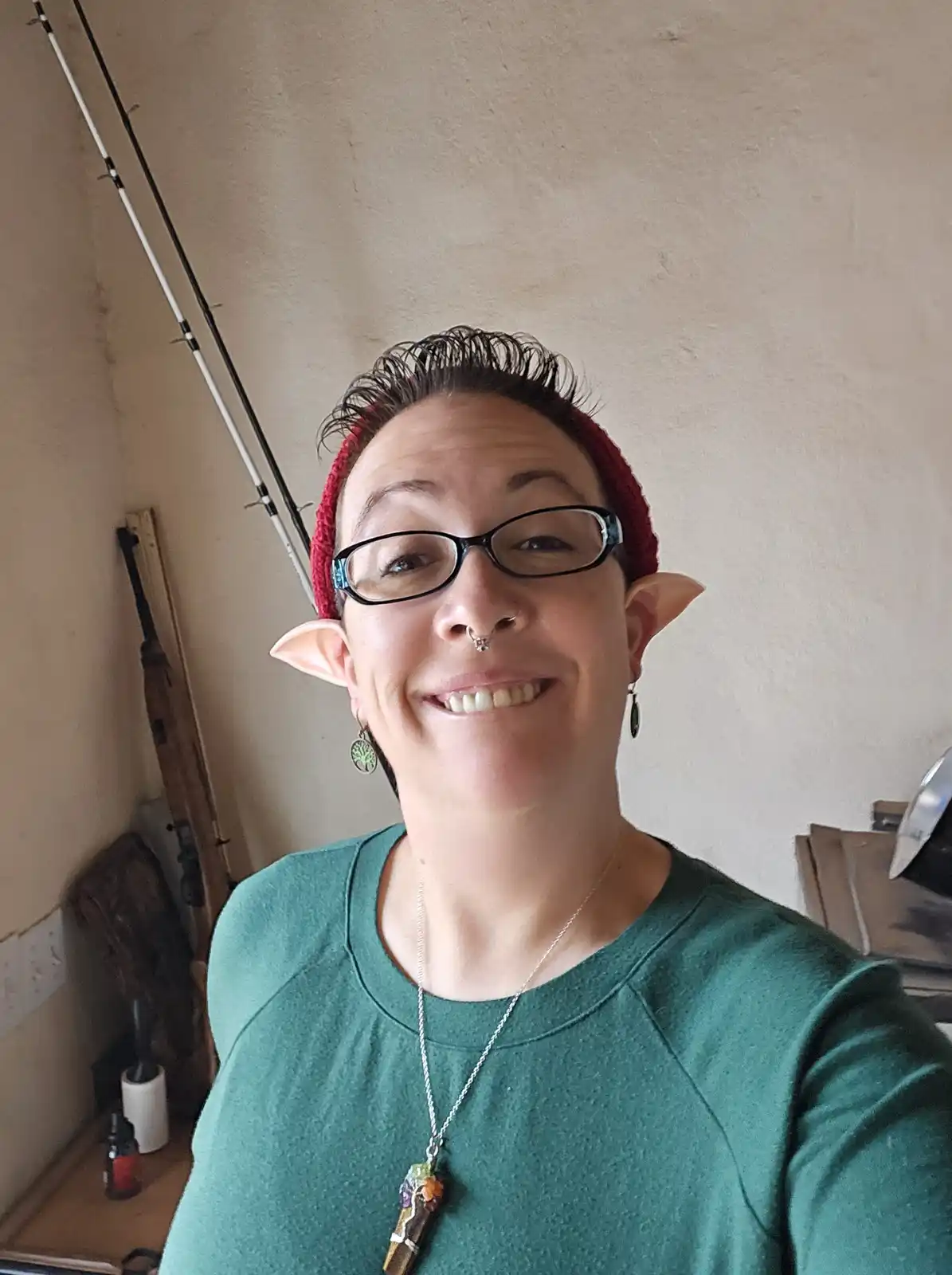 A smiling person with short dark hair, wearing a red beanie, glasses, and fake elf ears, poses for a selfie in a room with…