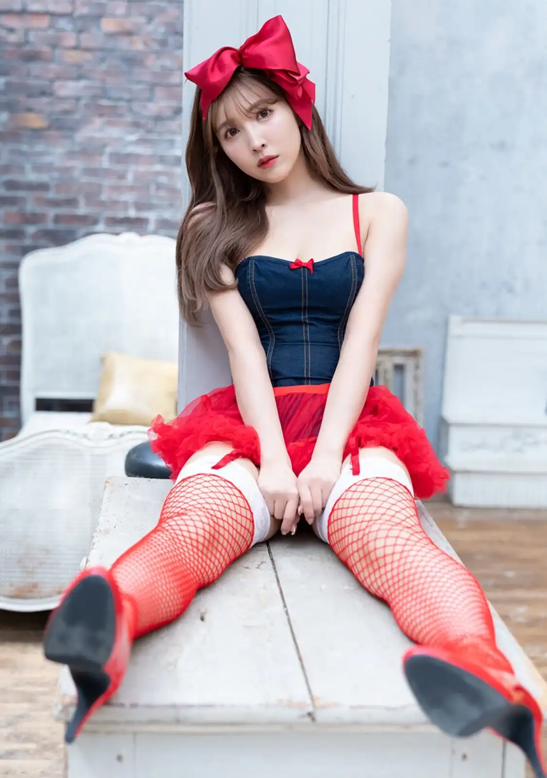 a young woman with long brown hair and a large red bow on her head sits on a white wooden bench. She wears a denim corset…