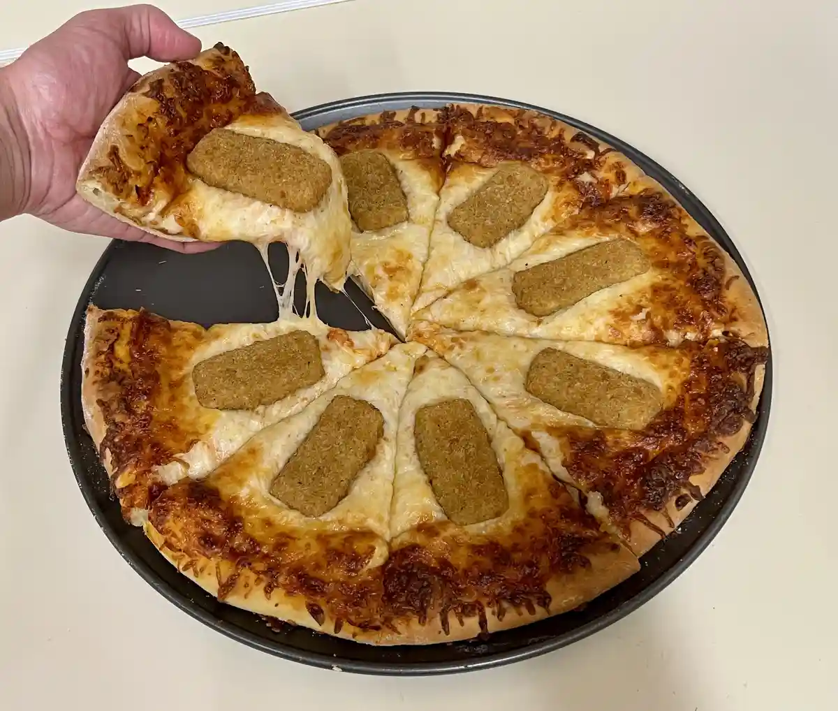 A hand lifts a slice of pizza from a black baking tray, showing melted cheese stretching between the slice and the rest of…
