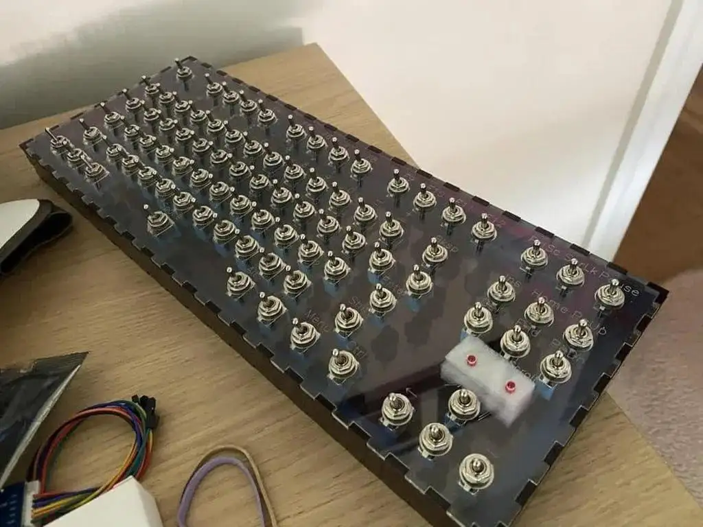 A custom-built keyboard with 100 silver toggle switches arranged in a grid on a dark, transparent surface sits on a wooden…