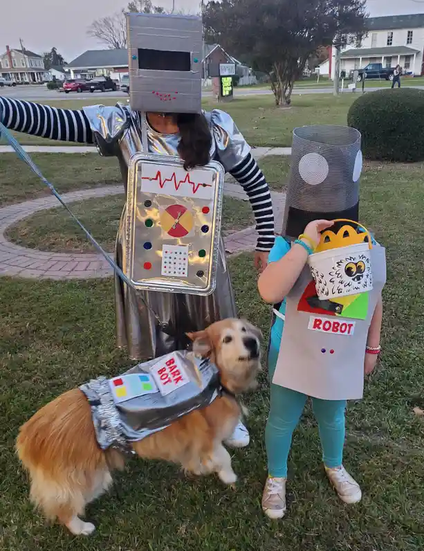 Two children dressed as robots stand on a grassy lawn, one wearing a silver foil costume with a cardboard head and control…