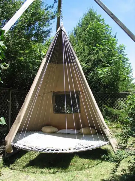 A beige teepee-style canopy hangs from a wooden frame in a backyard, suspended over a circular woven hammock bed with three…
