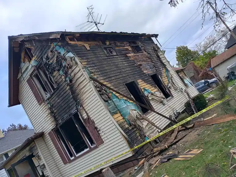 A house is severely damaged by fire, with charred siding, broken windows, and debris scattered around. Yellow caution tape…