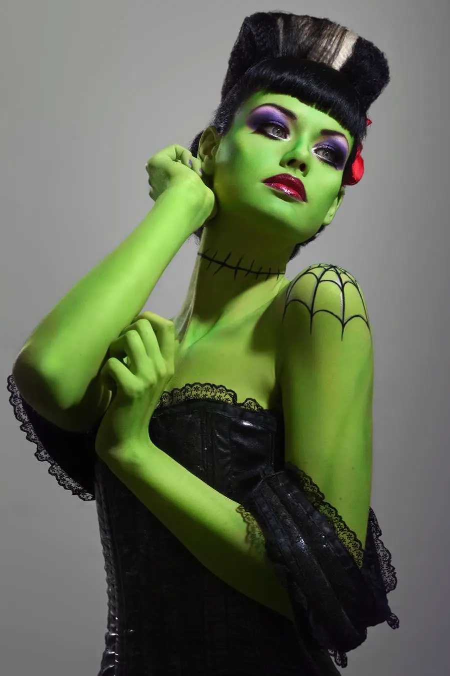 A person with green skin, black hair in a tall updo, and purple eye makeup poses in a black corset. Their neck has black…