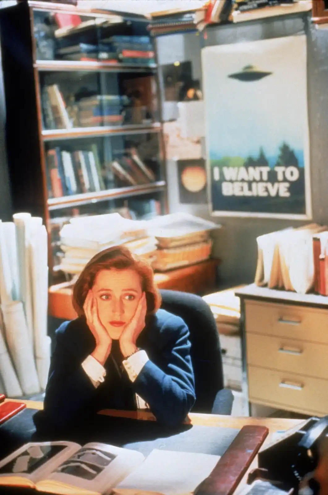 A woman sits at a cluttered desk in an office, hands on her cheeks, looking exhausted or overwhelmed. Behind her, a poster…