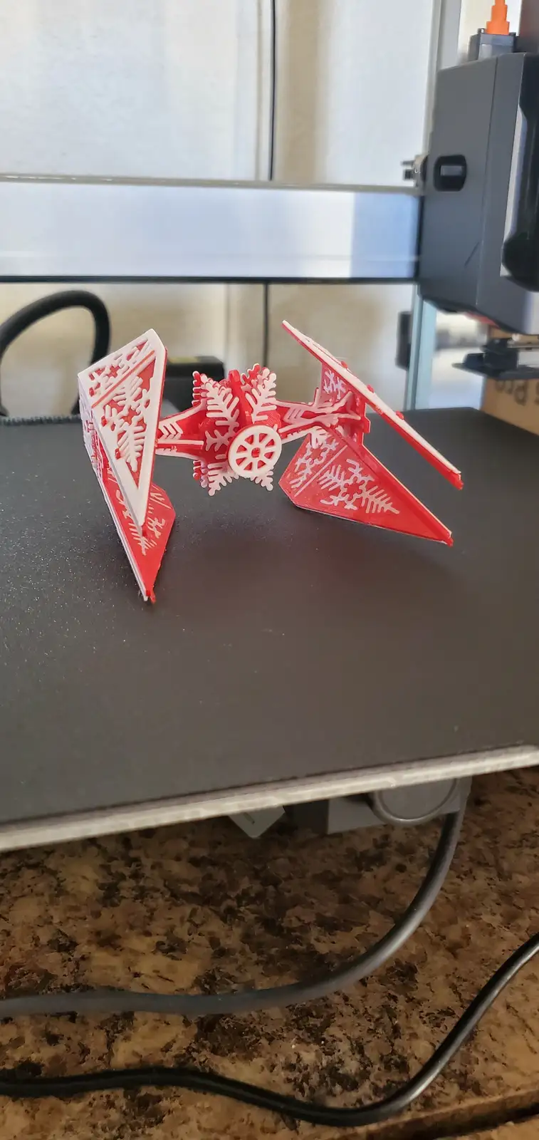 A red and white 3D-printed model of a TIE fighter from Star Wars, decorated with intricate snowflake patterns, sits on the…