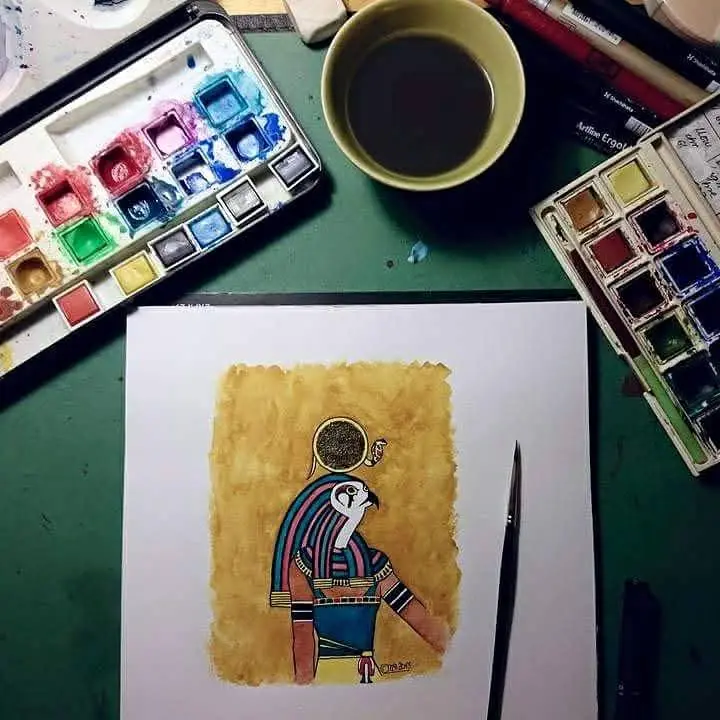 An overhead shot of an artist's workspace with watercolor palettes, a cup of coffee, and a painting of an Egyptian-style…