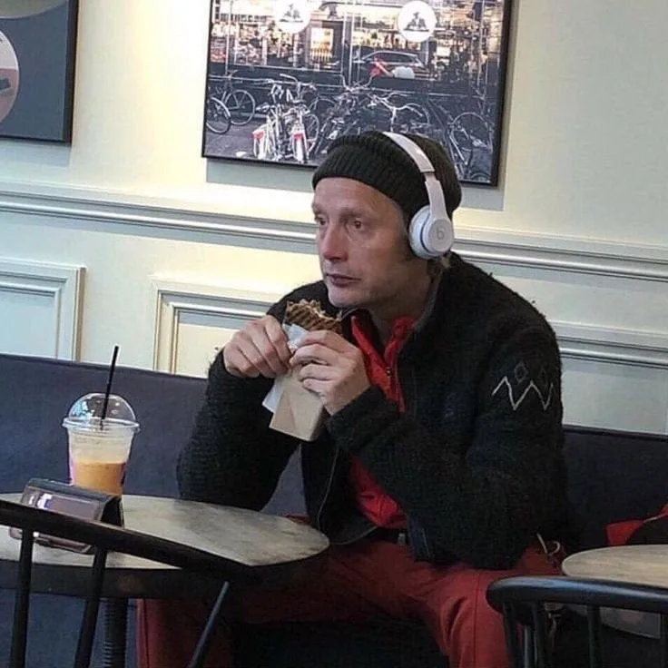 A man wearing white headphones and a black beanie sits at a cafe table, holding a waffle sandwich and looking surprised. A…