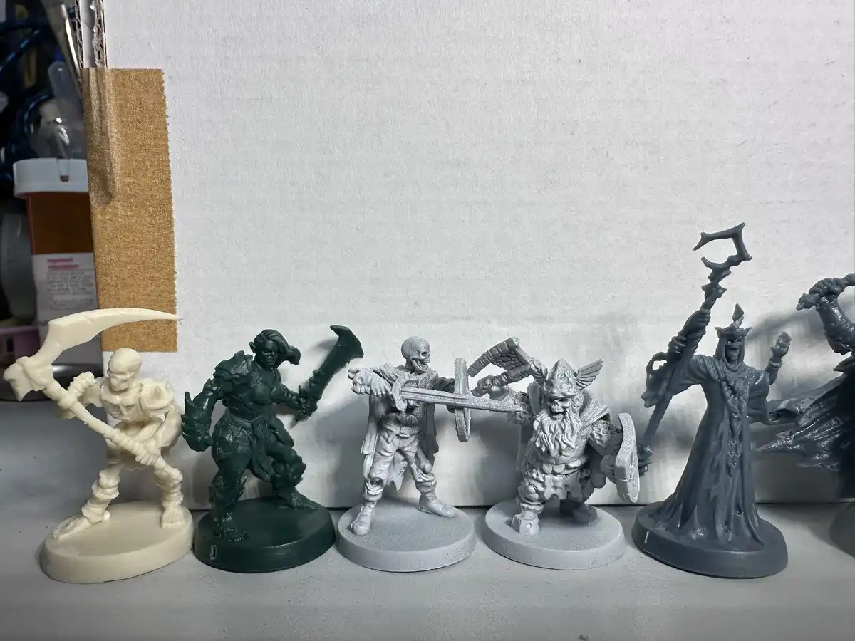 A row of unpainted fantasy miniatures, mostly in grey or white, stand on a shelf against a white wall. The figures include a…