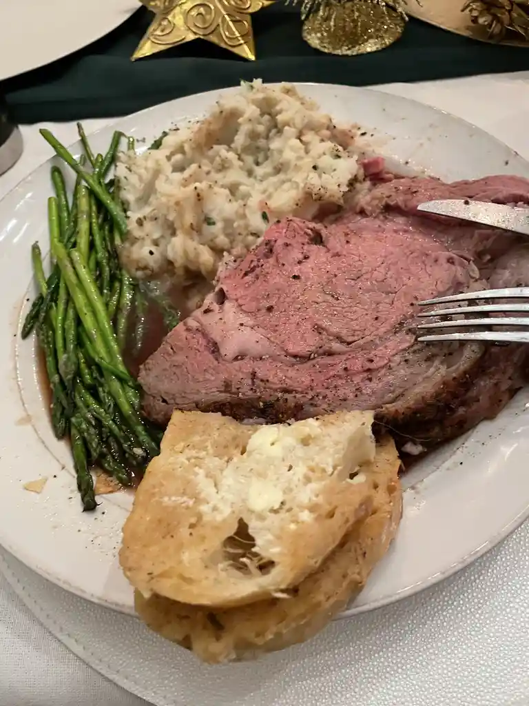 A close-up photo of a plated meal featuring a large slice of pink, medium-rare roast beef, mashed potatoes, green asparagus…
