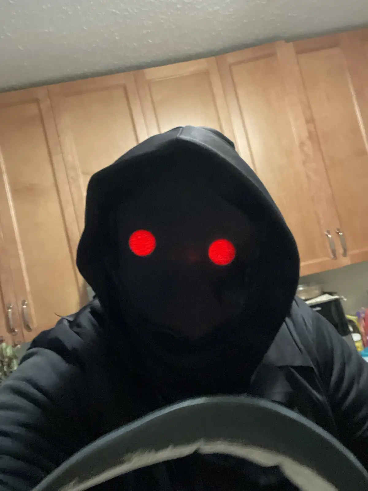 A person wearing a black hoodie with glowing red LED eyes in the hood, taking a selfie in a kitchen with wooden cabinets…