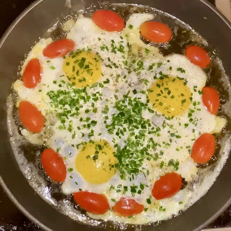 A close-up photo shows a frying pan containing several sunny-side-up eggs arranged in a circular pattern, surrounded by…