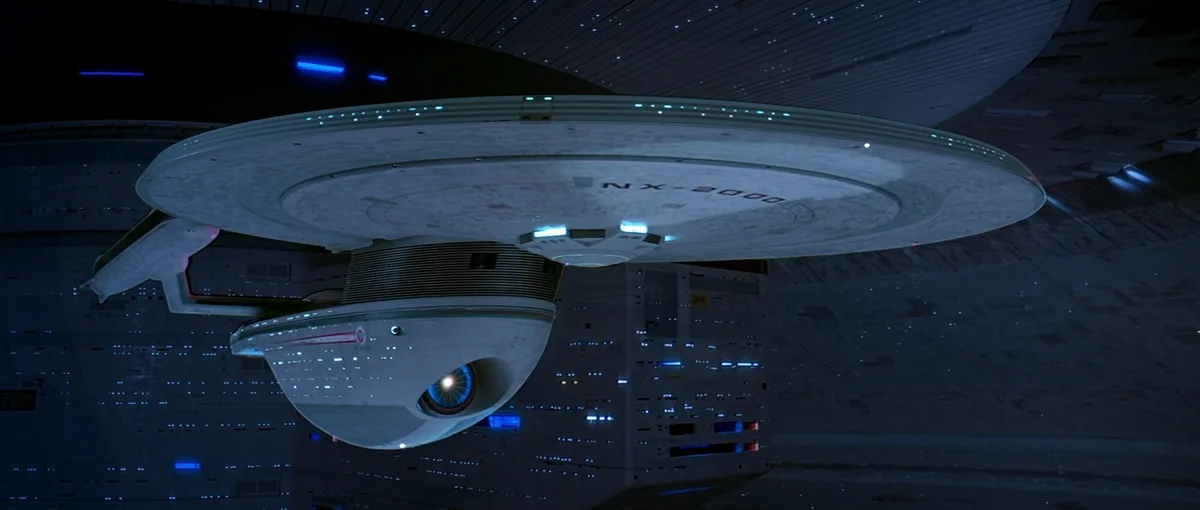A Star Trek Enterprise NX-01 spaceship is shown docked inside a futuristic space station, with blue lights illuminating its…
