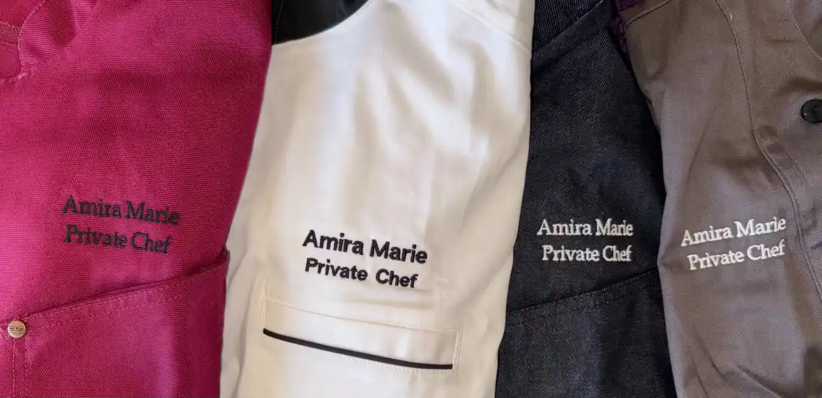 A close-up shot displays four different colored chef jackets—pink, white, black, and gray—each with the same embroidered…