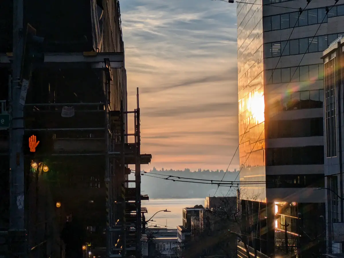 A city street view at sunset framed by two tall buildings, one with scaffolding on the left and a glass skyscraper on the…