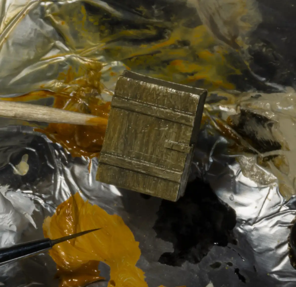 A close-up photo of a painter's palette covered in crumpled aluminum foil, smeared with thick globs of yellow, black, and…