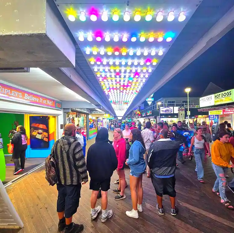 A crowded boardwalk at night with people walking under a large, colorful light installation shaped like a heart. The lights…