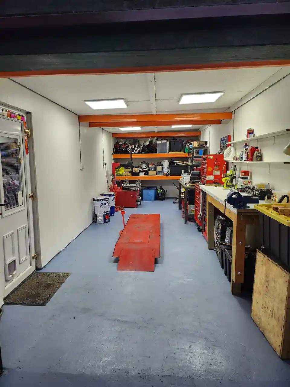 A well-lit, tidy garage with blue flooring, orange beams, and white walls, featuring shelves with helmets and tools…