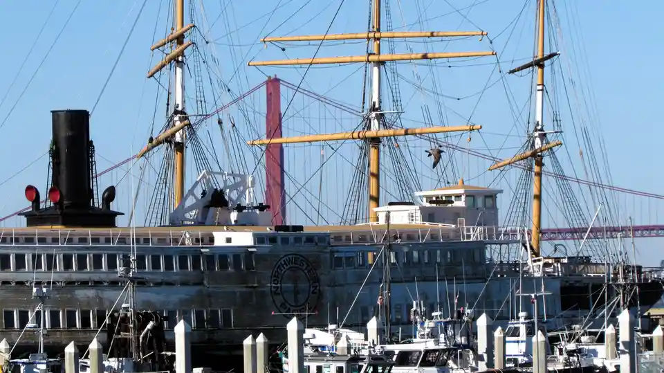 A large, weathered ship with multiple tall masts and rigging is docked in a harbor, with a red suspension bridge visible in…