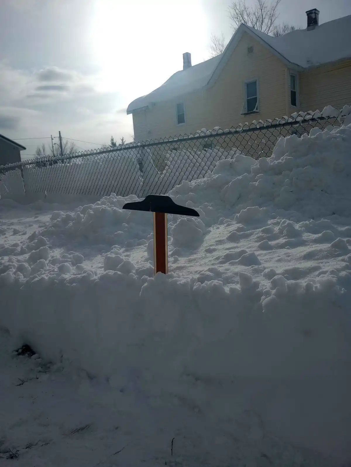 A snow shovel is stuck upright in a large pile of snow, with a yellow house and chain-link fence visible in the background…