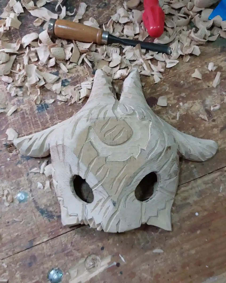 A partially carved wooden owl mask lies on a workbench covered in wood shavings, with carving tools nearby. The mask has two…