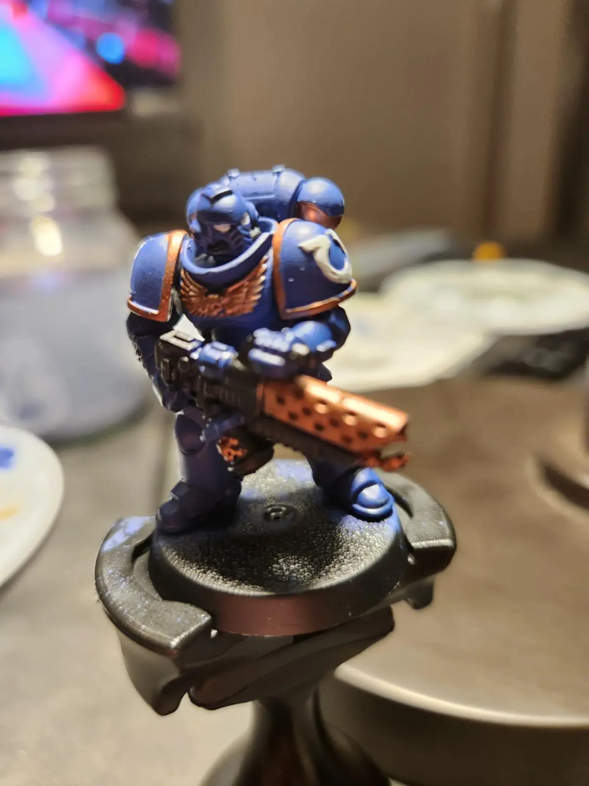 A close-up photo of a blue and orange painted miniature of a Space Marine from Warhammer 40,000, standing on a black base…