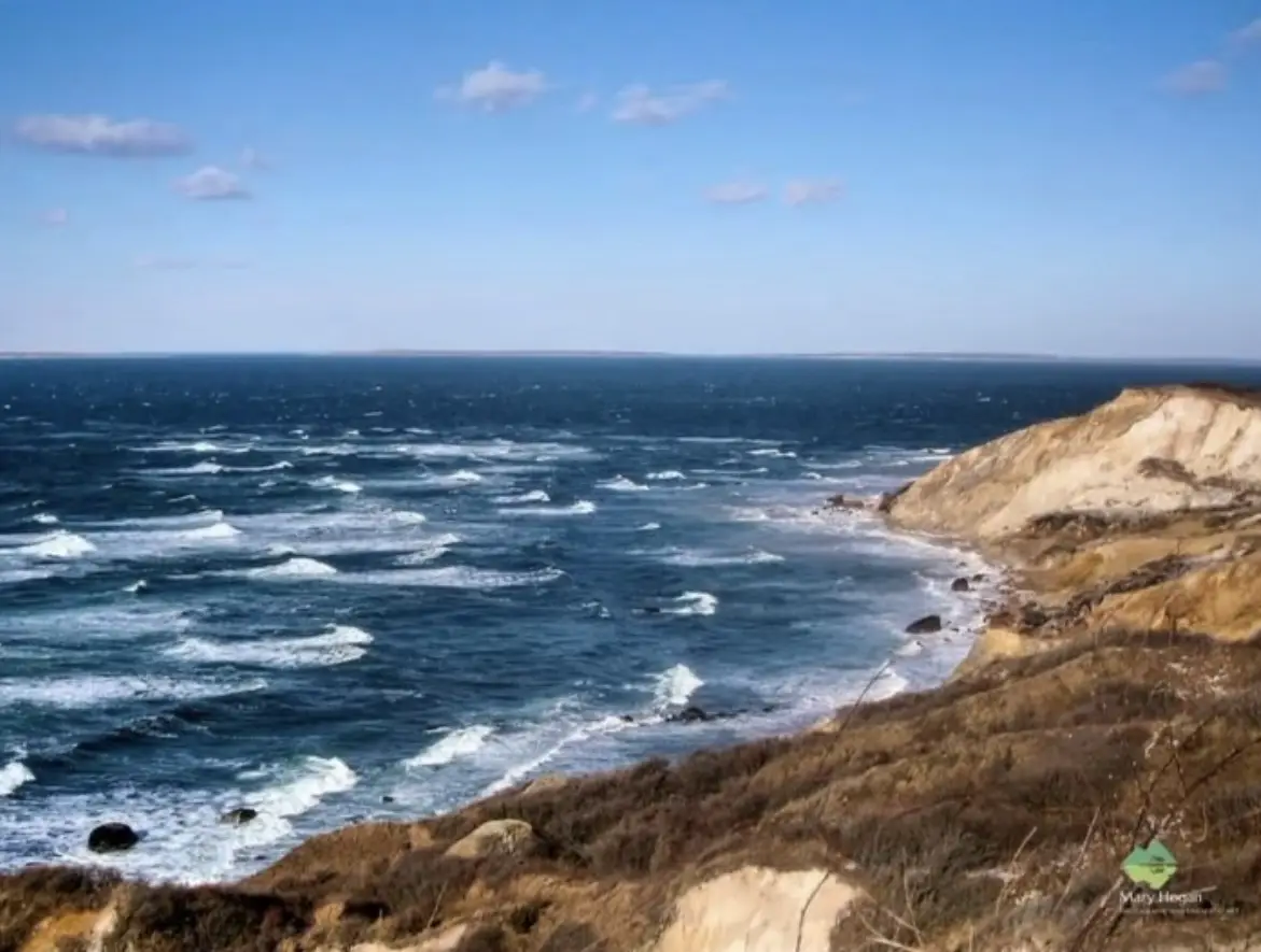A scenic photo of a choppy ocean with waves crashing against a sandy, grassy cliffside under a partly cloudy blue sky. The…