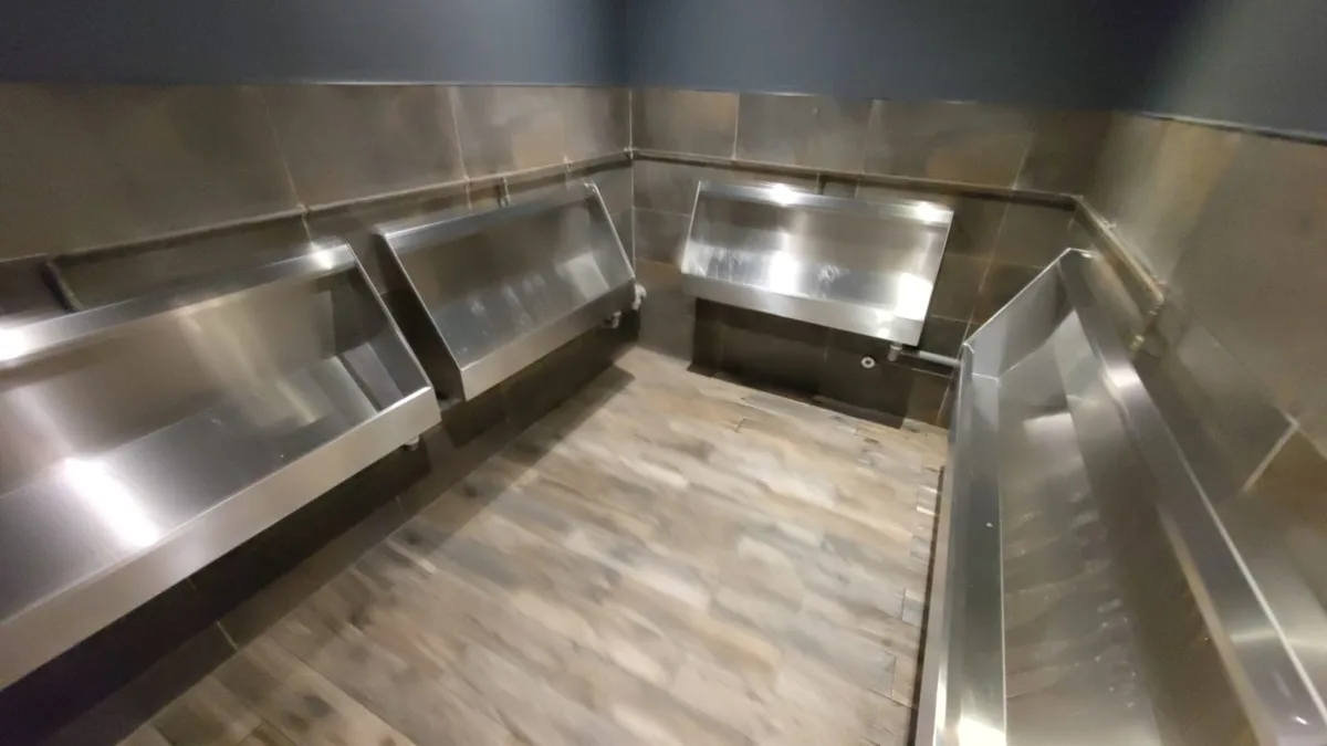 A photo of a modern restroom with four stainless steel urinals mounted on tiled walls, set against a wood-patterned floor…