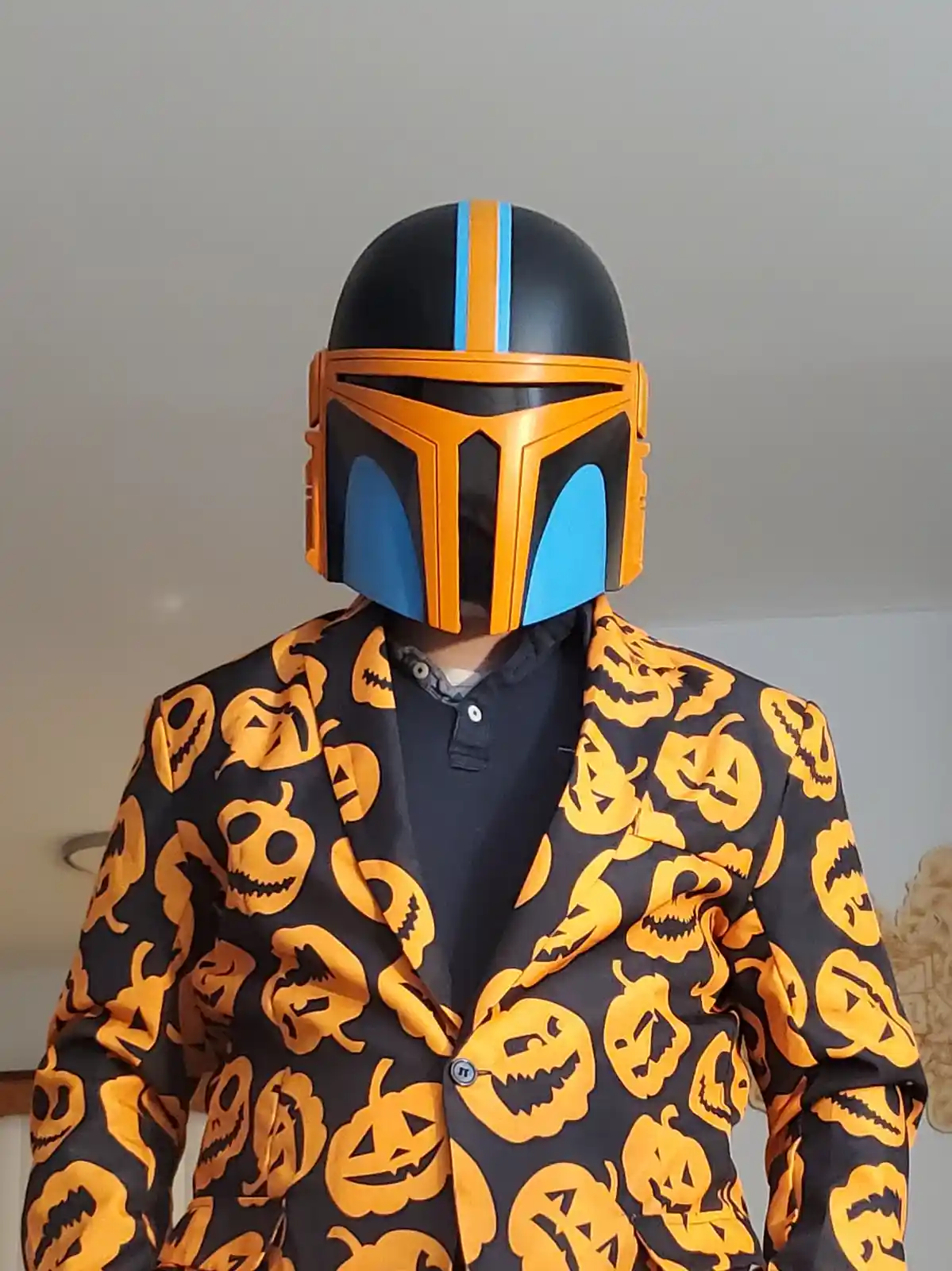 A person wearing a Mandalorian-style helmet with orange and blue accents, paired with a black blazer covered in orange…