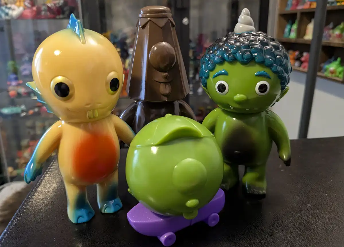 Four colorful, stylized toy figures are arranged on a black surface. A yellow figure with blue fins stands left, a brown…