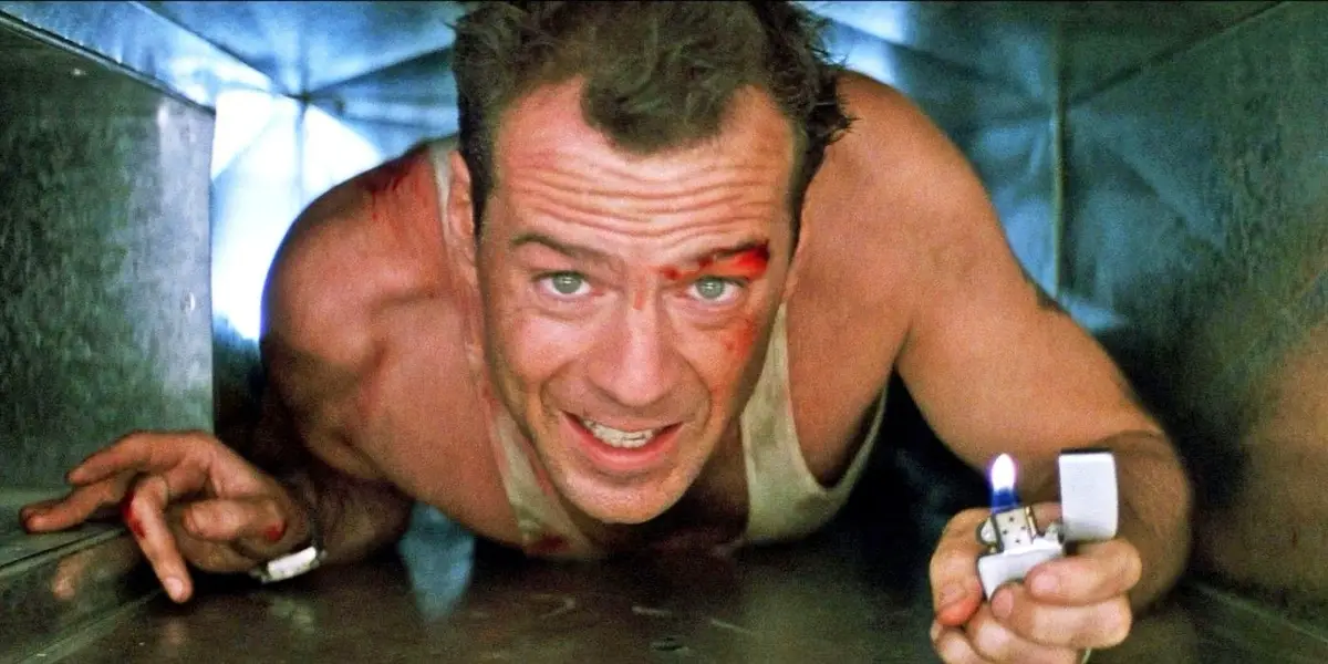 A shirtless man with blood on his face and a white tank top is crawling through a metal duct, holding a lit lighter in his…