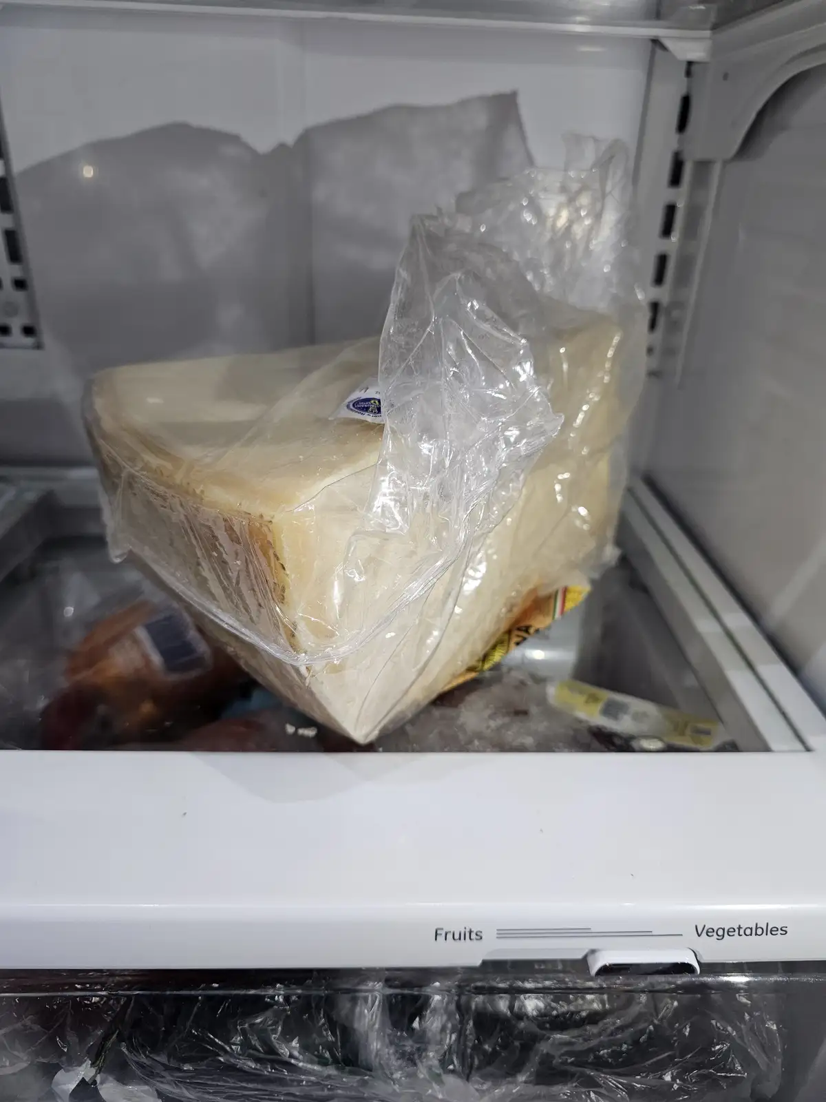 A large wedge of cheese wrapped in plastic sits inside a freezer compartment labeled 'Fruits' and 'Vegetables', with other…