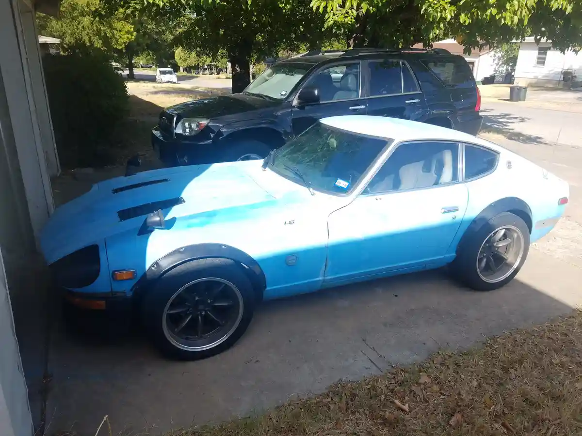 A light blue, weathered Datsun 240Z sports car with black fender flares and aftermarket wheels is parked in a driveway next…