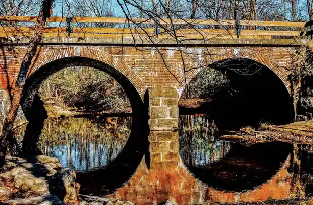 A scenic photo of a brick arch bridge over a calm river, with its reflection perfectly mirrored in the water below. Bare…