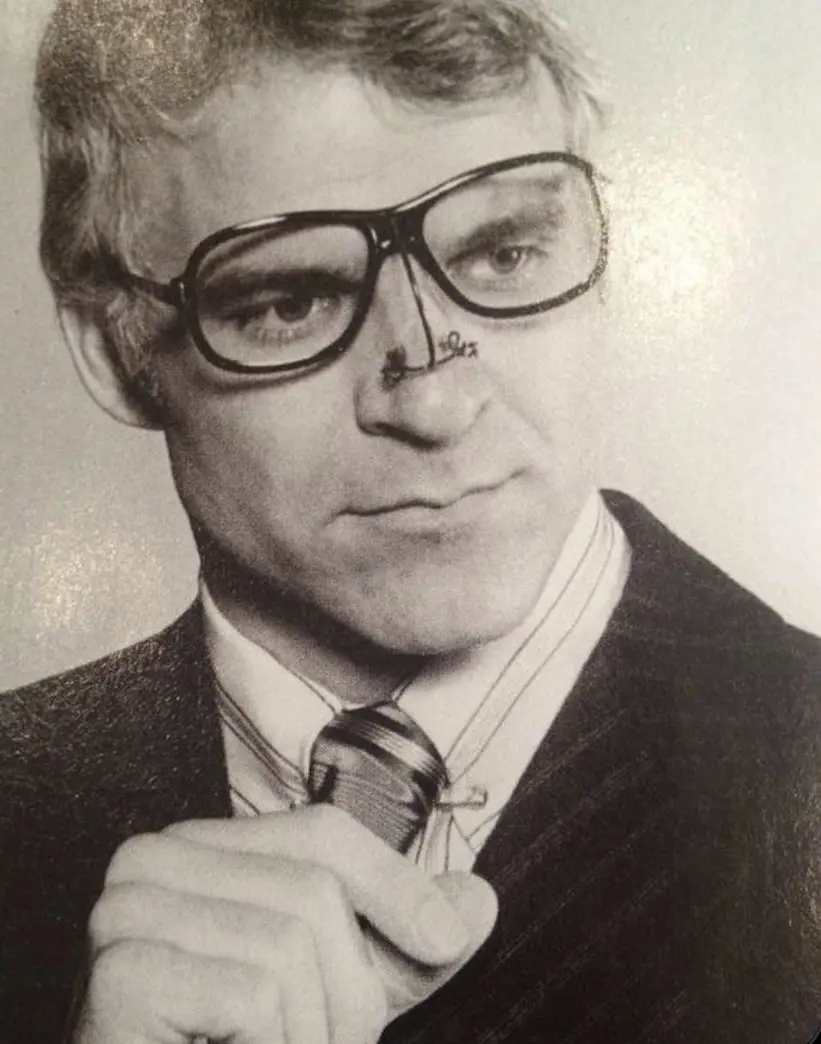 A black-and-white photo of a man with thick-rimmed glasses and a pinstripe suit, looking directly at the camera with a…
