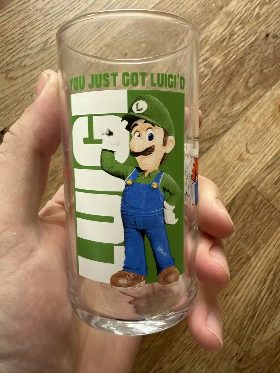 A hand holds a clear drinking glass against a wooden surface. The glass features a printed design of the video game…