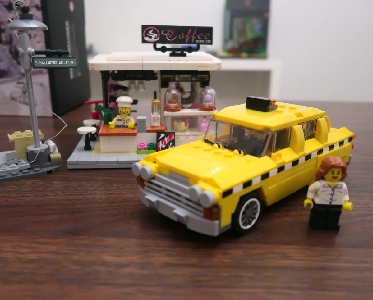 A LEGO diorama shows a yellow taxi and a female LEGO minifigure standing beside it, with a coffee shop in the background…