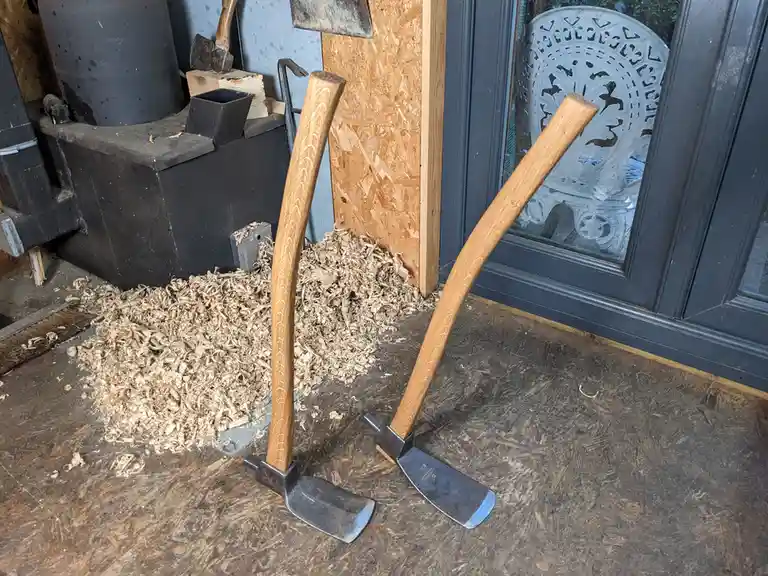Two wooden-handled axes lean against a wall in a workshop, surrounded by wood shavings on an OSB floor. A black wood stove…