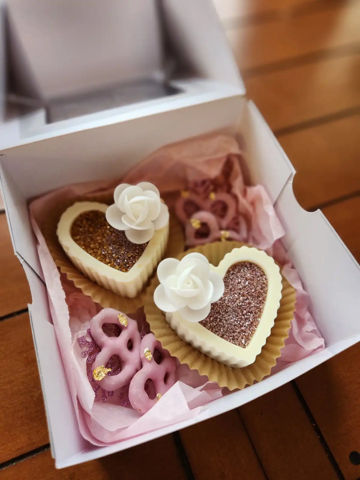 A white box lined with pink tissue paper contains heart-shaped pastries with white icing roses and glittery centers…