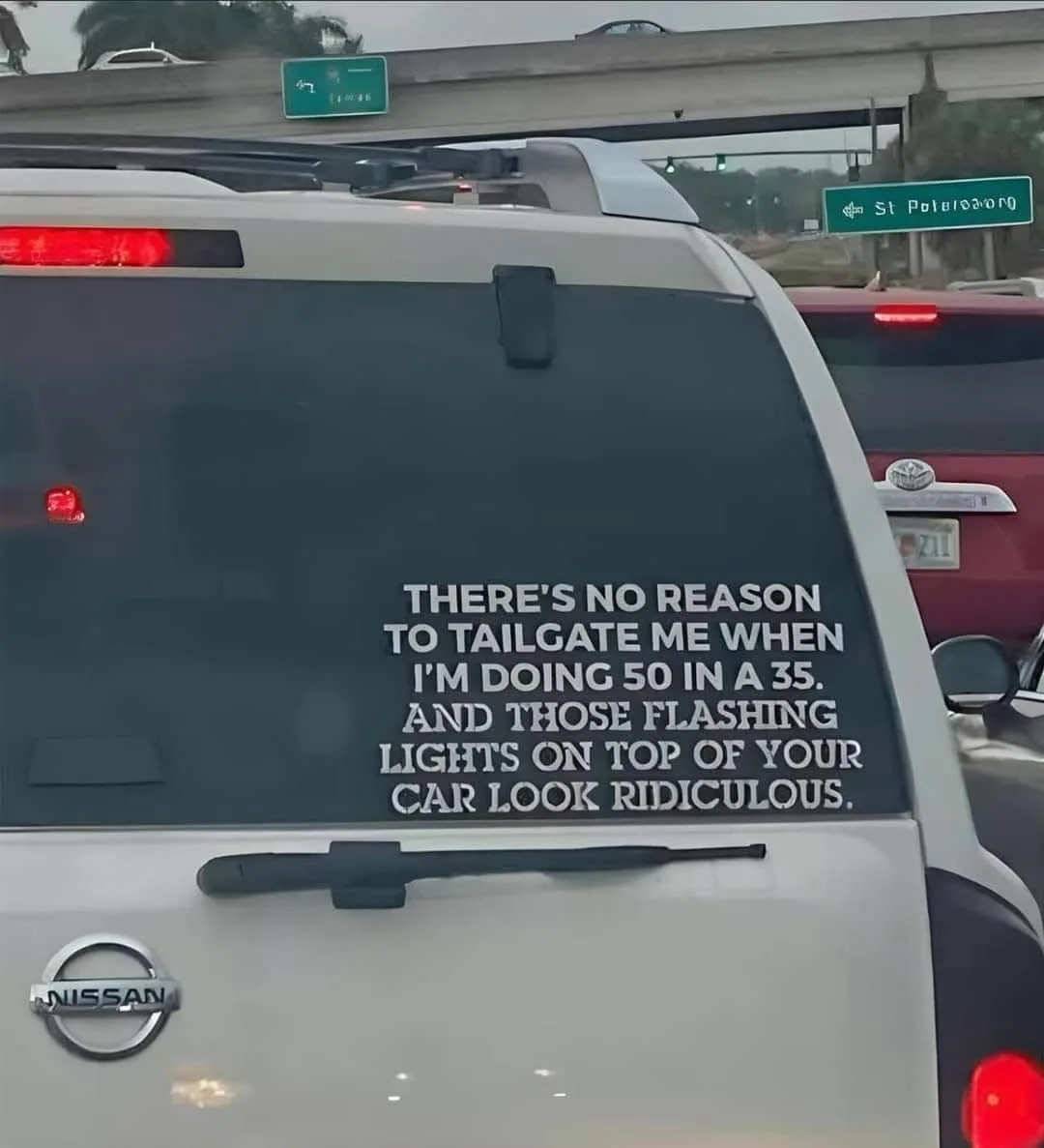 The image shows the rear of a white Nissan SUV on a highway, with a large white decal on the back window. The decal reads…