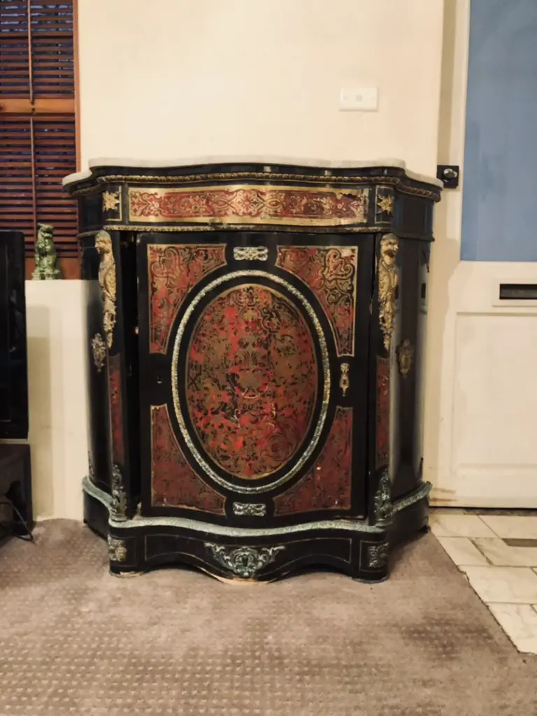 a black and red ornate cabinet with gold detailing stands in a room with carpet and a white door, overlaid with the text…