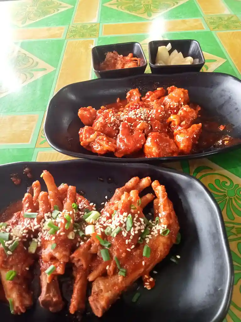 Two black plates of Korean-style spicy chicken dishes sit on a green and yellow patterned tablecloth. The top plate holds…