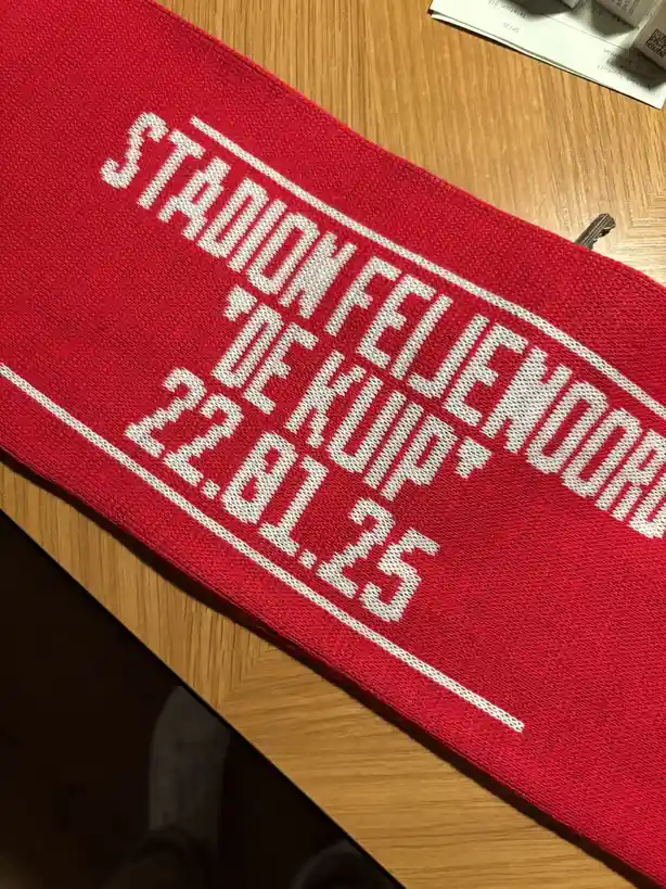 A red knitted scarf with white text reading 'STADION FEIJENHOOCH 'DE KUIP' 22.01.25' lies diagonally on a wooden table, with…