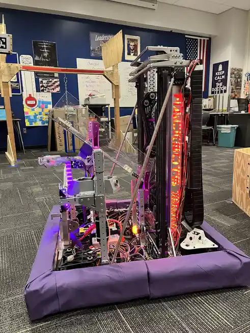 A complex, custom-built robot with exposed wiring, metal frames, and purple and red LED lights sits on a purple padded base…