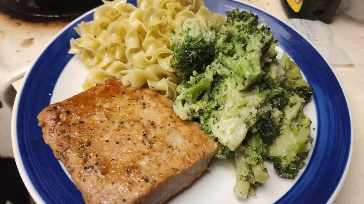 A close-up photo shows a white plate with a blue rim holding a cooked salmon fillet, a serving of pasta, and a large portion…