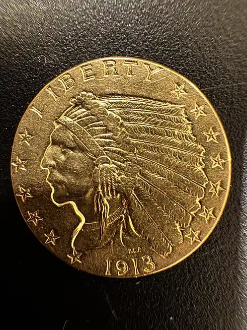 A close-up photo of a gold-colored 1913 Indian Head $2.50 gold coin, showing the profile of a Native American wearing a…