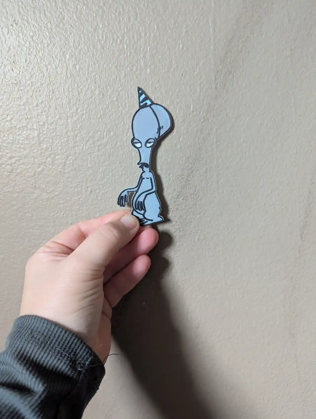 A hand holds a light blue, 3D-printed cutout of the cartoon alien character from 'Invader Zim' wearing a party hat, against…