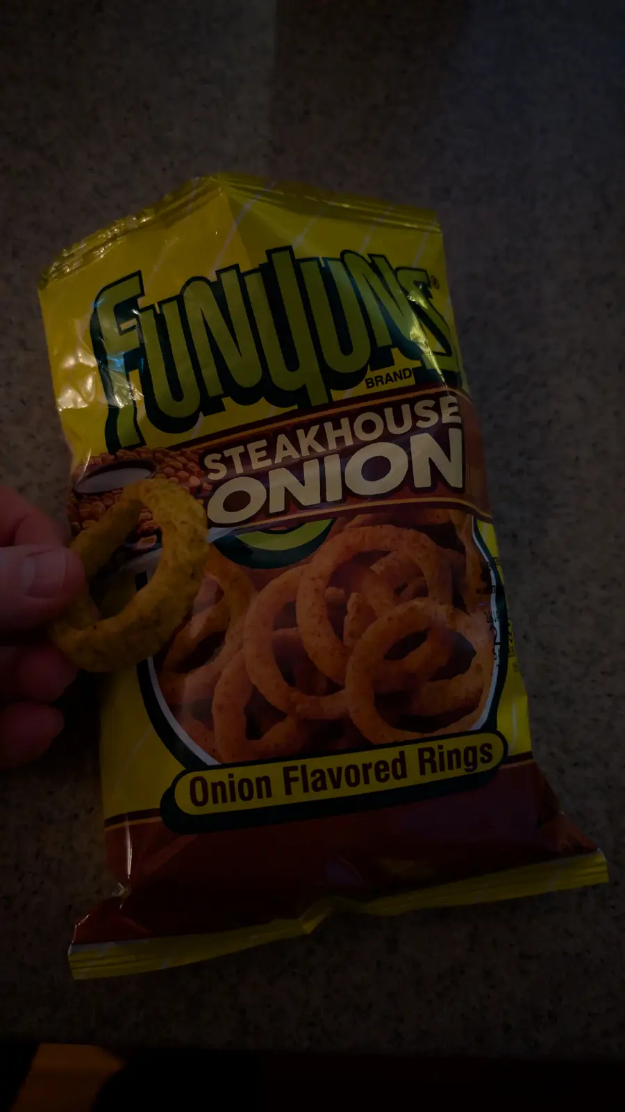 A hand holds a single Funjuns Steakhouse Onion flavored ring in front of its yellow and brown packaging, which clearly…