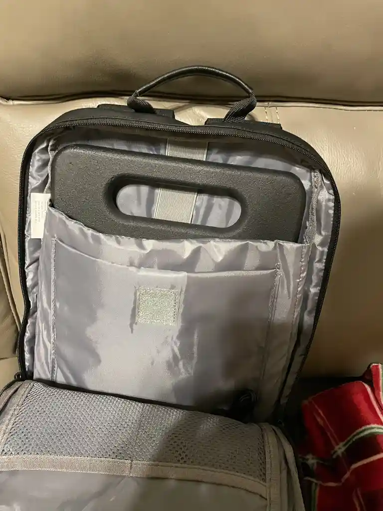 A black backpack is open on a beige leather couch, revealing a gray interior with a large, black, foam-like handle insert…