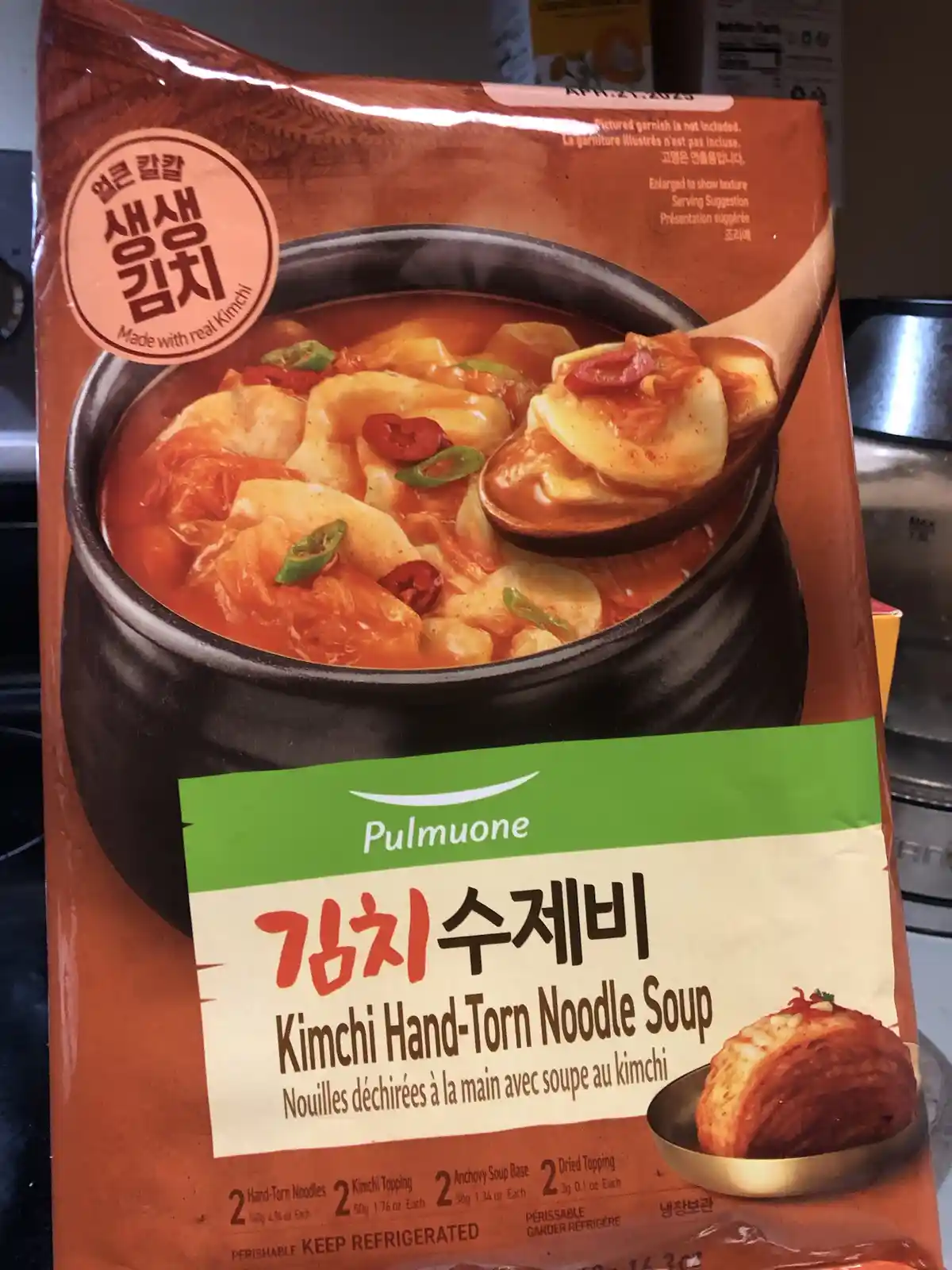 A close-up photo of a Pulmuone brand kimchi noodle soup package, featuring an image of a steaming bowl of red soup with…